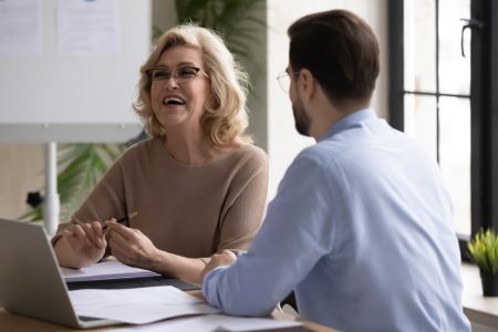 Overjoyed middle aged senior female leader having fun, enjoying informal conversation with young male colleague at office meeting. Happy mature mentor laughing at funny joke with partner indoors.